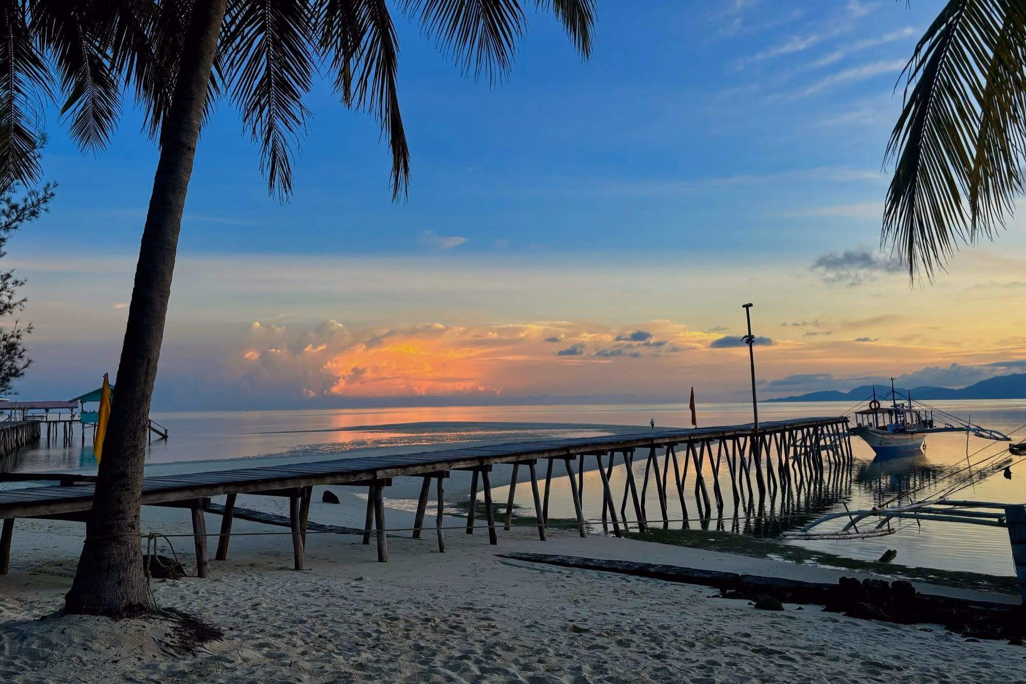 Sunset over the sea at Onuk Island in Balabac, with a wooden jetty on stilts, palm trees, white sand and an outrigger boat moored on the right.