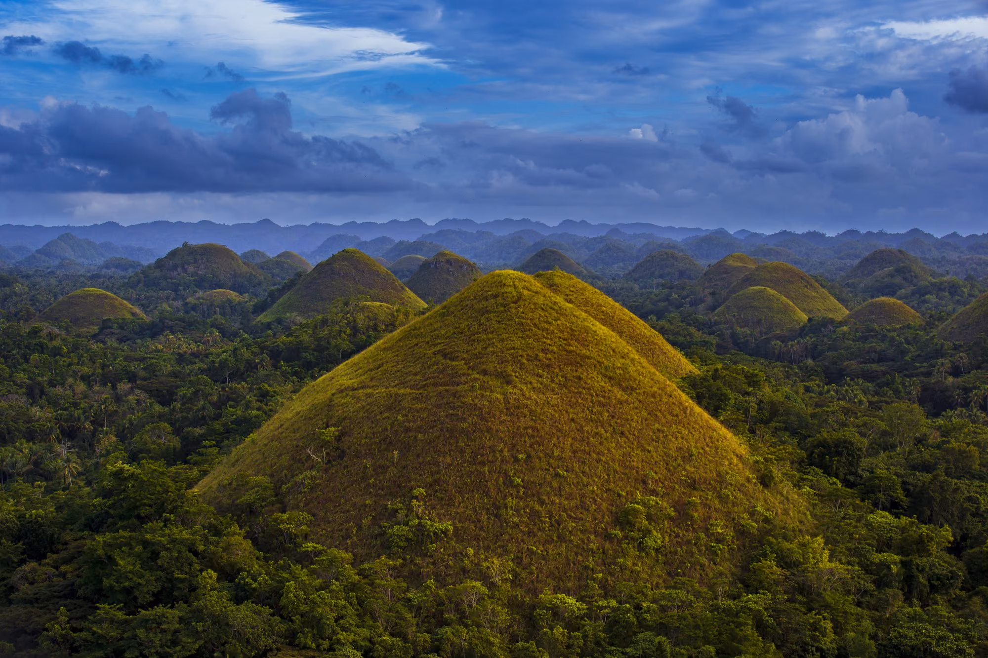 The cone-shaped Chocolate Hills of Bohol rising above green forest, under a blue sky with clouds.