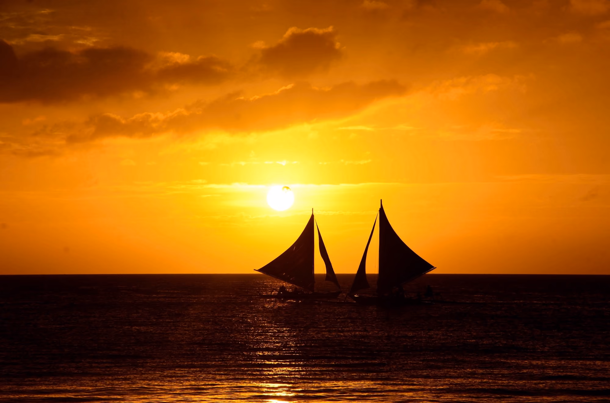 Two paraw sailboats in silhouette against a golden sunset over the sea off Boracay.