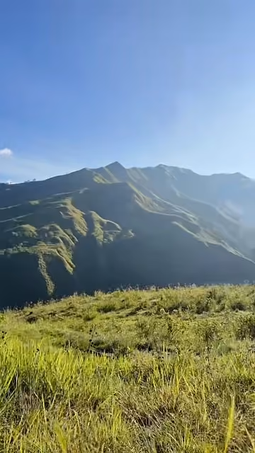Green grass on a hillside in Bukidnon, with the mountain range behind it and a blue sky.