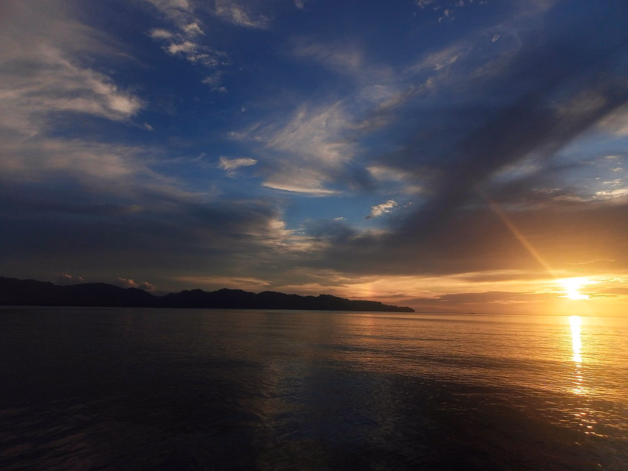 Illustrative photo of the sun setting over Dapitan Bay, with a golden reflection on the water and a dark headland on the horizon.