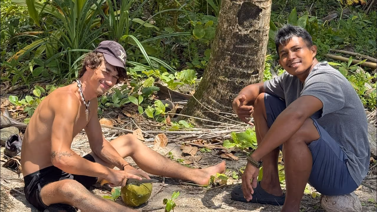 Robert sitting on the sand opening a coconut, next to a man from the island, under the palms at the top of a Dinagat beach.