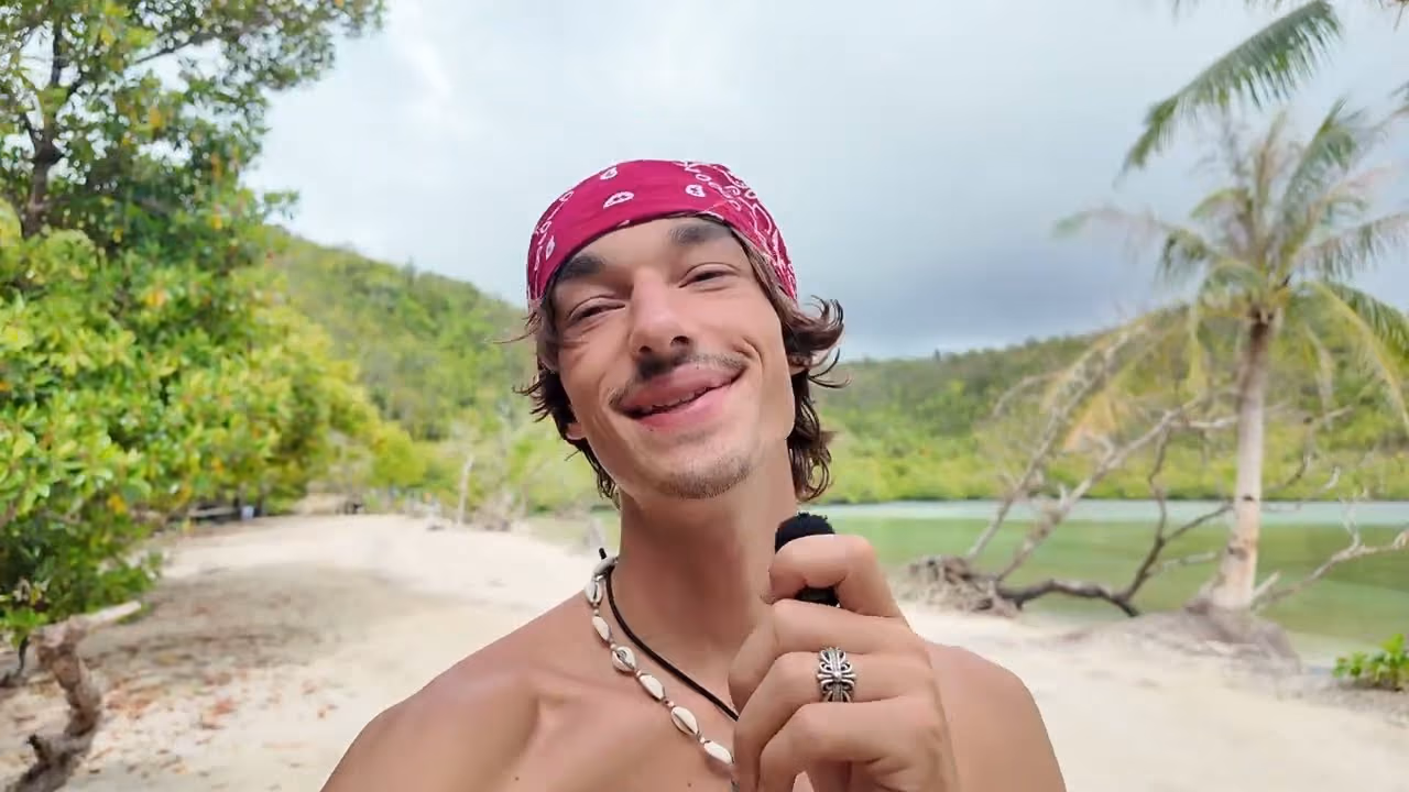 Robert on a beach on Dinagat, talking into the camera, with a green lagoon and palms behind him.