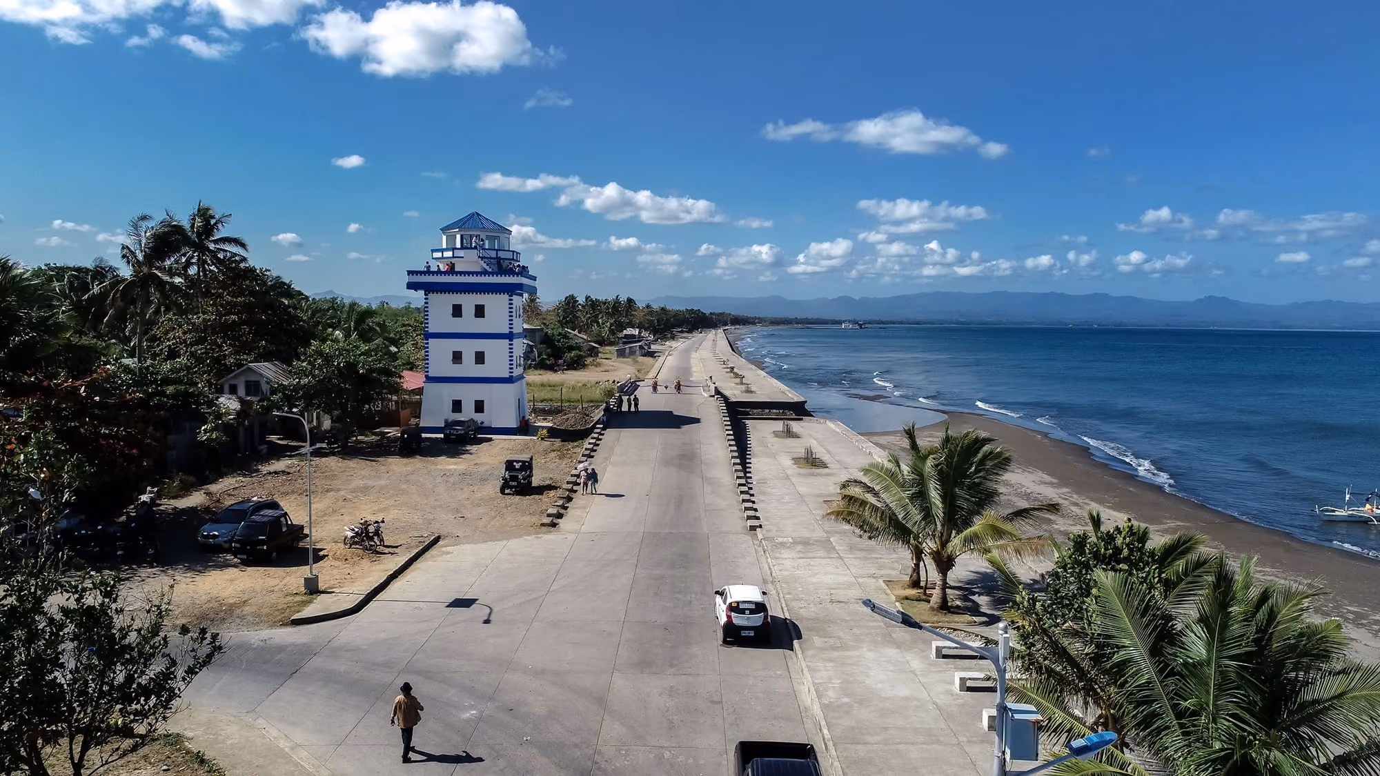Illustrative photo of the Dipolog City Boulevard from above: the seaside road, a white lighthouse tower, palms and a dark-sand beach along a blue sea.