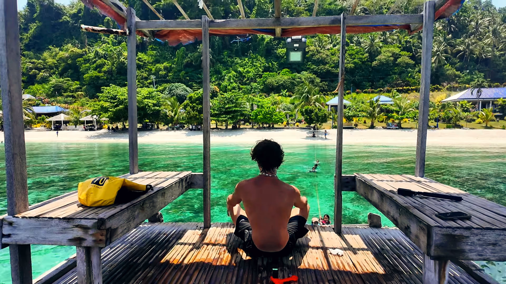 Robert sitting on a wooden deck over clear water, looking out at a palm-lined beach.