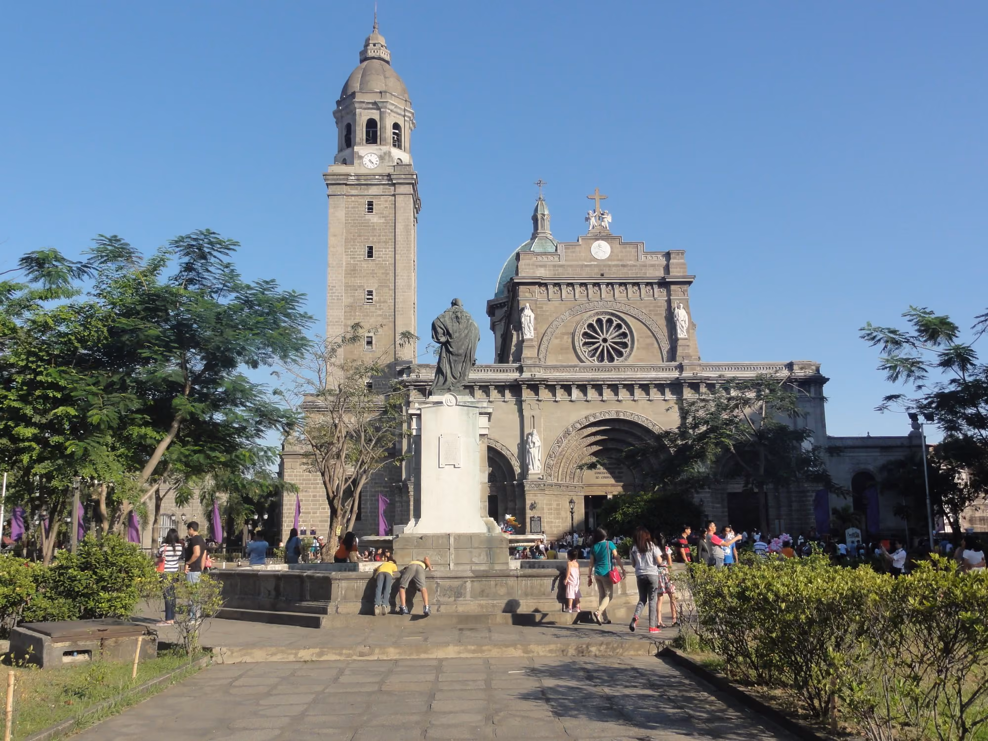 Manila Cathedral on Plaza Roma in Intramuros, Manila, in warm afternoon sun under a clear blue sky, with trees and visitors in the square.