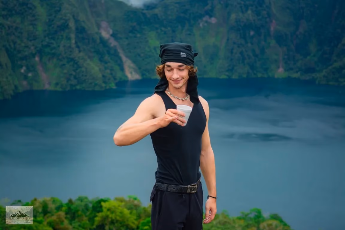 Robert in a black vest and bandana lifting a cup, with the dark blue Lake Holon and the green crater wall behind him.