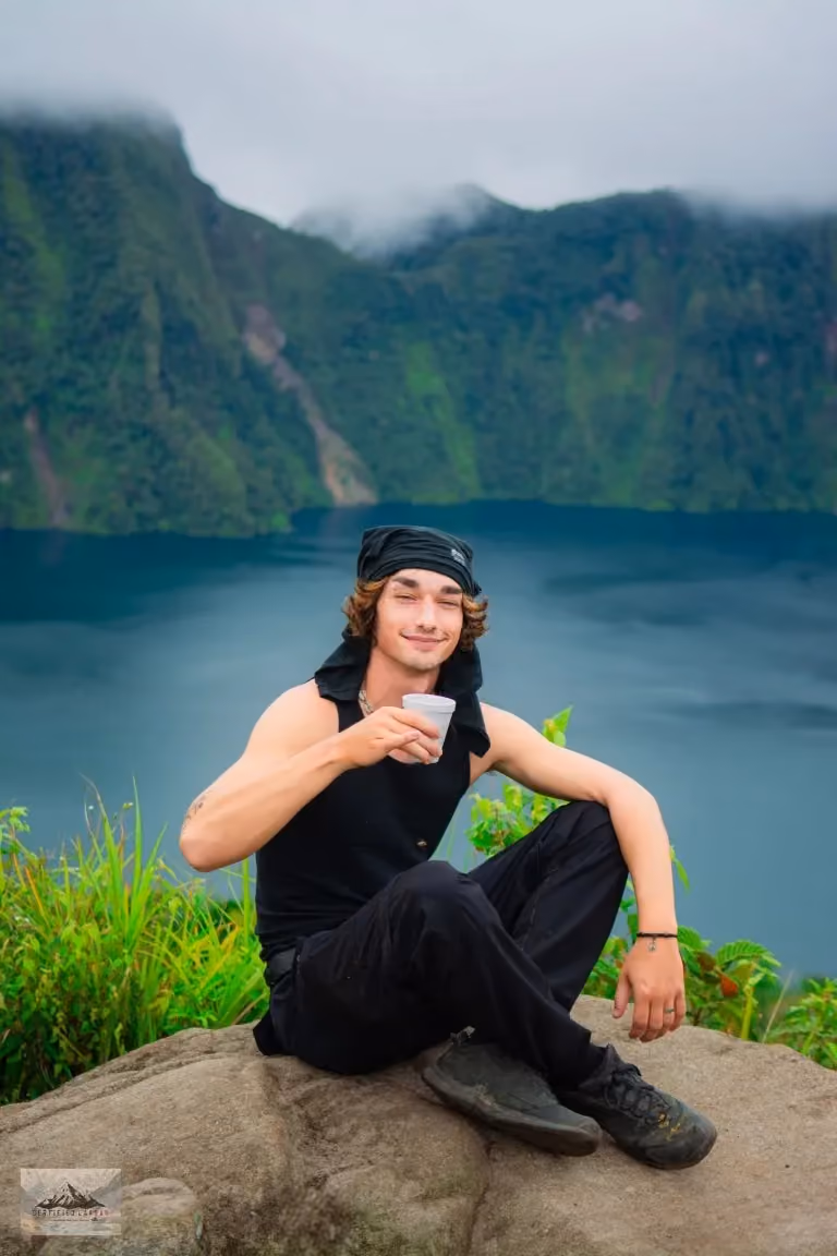 Robert in a black vest and bandana sitting on a rock at the edge of a cliff, drinking from a cup, with Lake Holon and the forested crater below him.