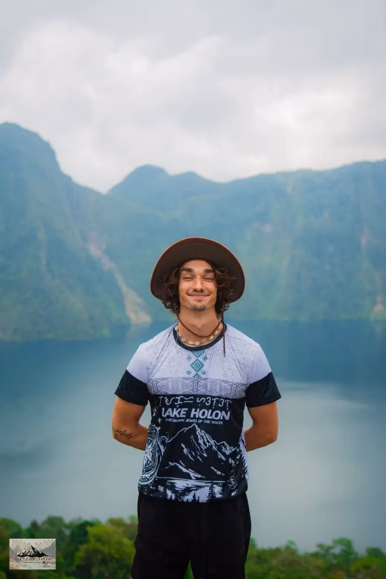Robert in a hat and a Lake Holon t-shirt, standing in front of the blue crater lake with green mountain walls rising behind him.