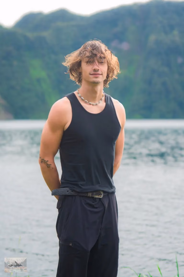 Robert in a black vest standing at the edge of Lake Holon, with a necklace on and the green crater wall behind him.