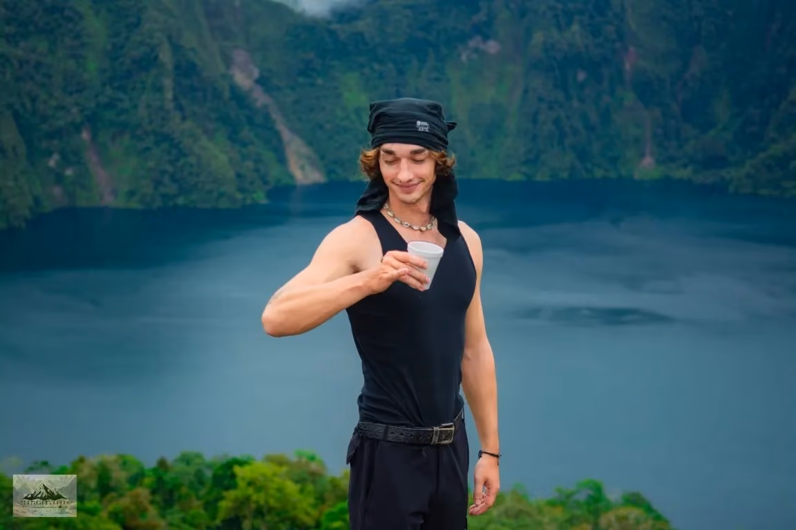 Robert sitting on a rock above the dark blue Lake Holon with a cup in his hand, green crater walls behind him.