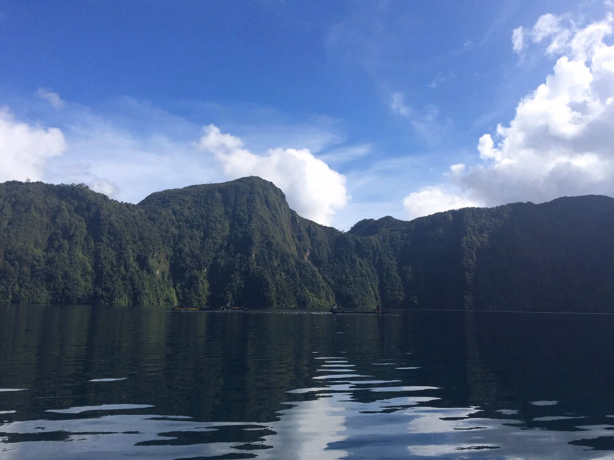 Lake Holon in South Cotabato from the water: forested crater walls under a blue sky with white clouds, their reflection on the calm lake, and two small boats near the far shore.