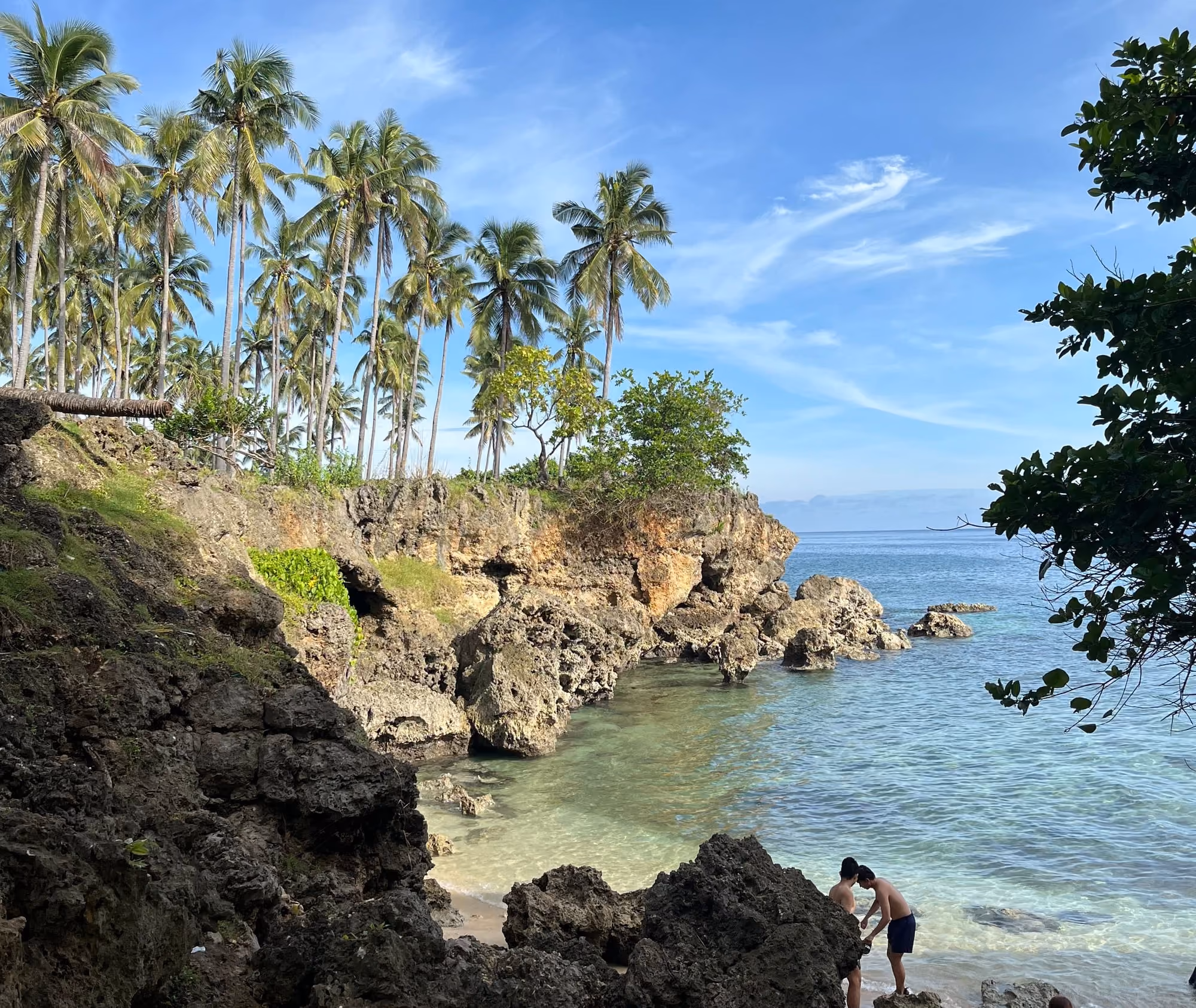 Illustrative photo of a small cove in Liloy, Zamboanga del Norte: coconut palms on a limestone cliff, clear turquoise water and a few swimmers on the rocks.