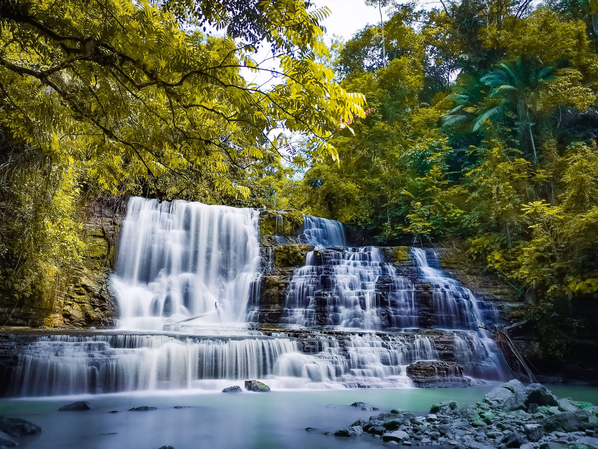 Illustrative photo of Merloquet Falls in Zamboanga City: a wide, two-tiered waterfall spilling over layered rock into a turquoise pool, with jungle on both sides.