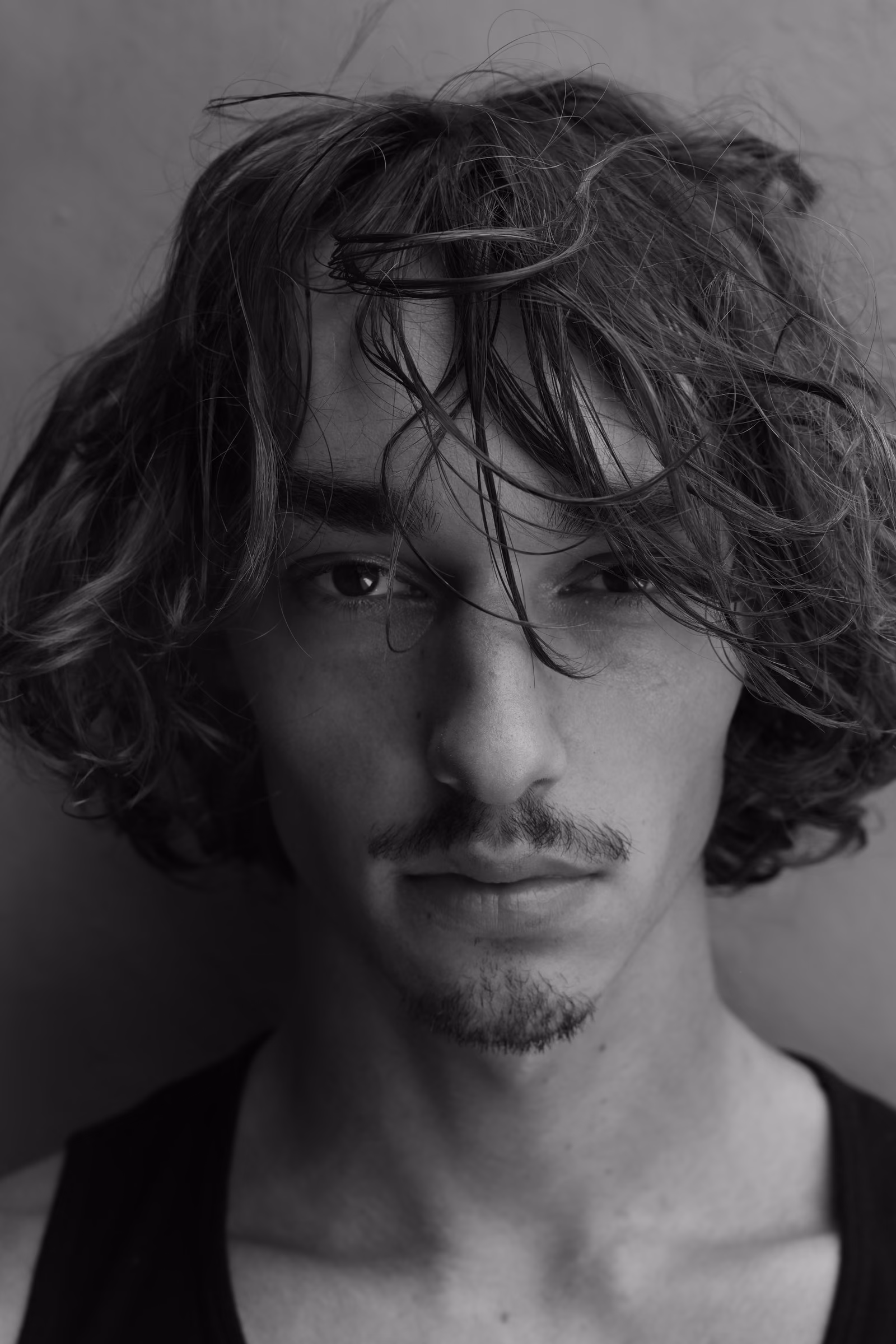 Black-and-white portrait of a man with tousled hair, looking straight into the camera against a plain grey wall.