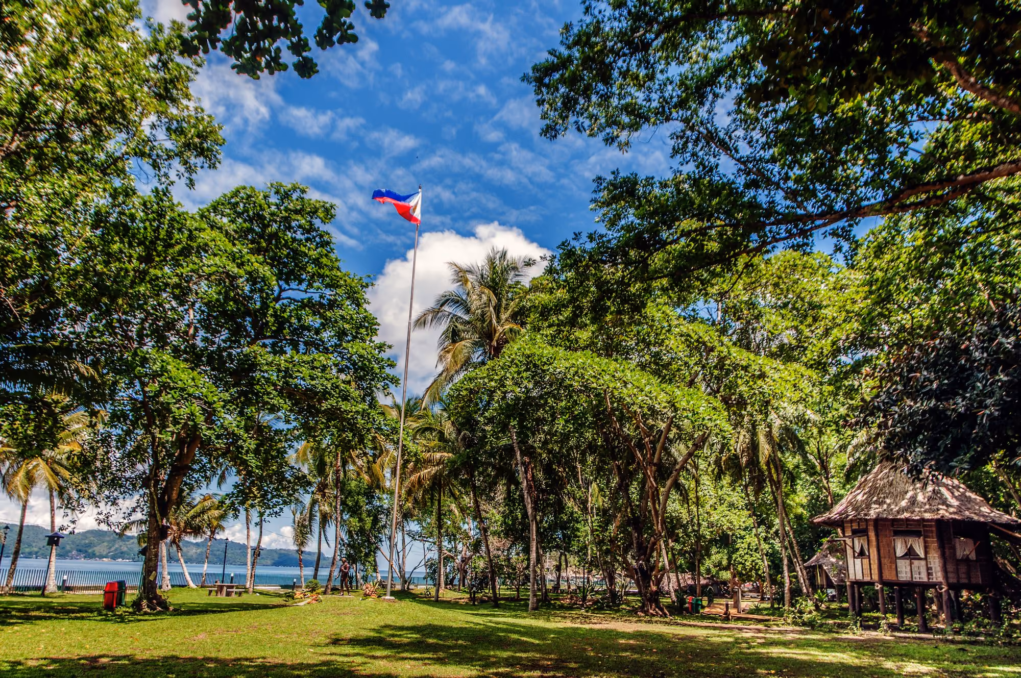 The Rizal Shrine park in Dapitan City: tall shade trees and palms on a green lawn, the Philippine flag against a blue sky, a nipa house on stilts and Dapitan Bay behind.