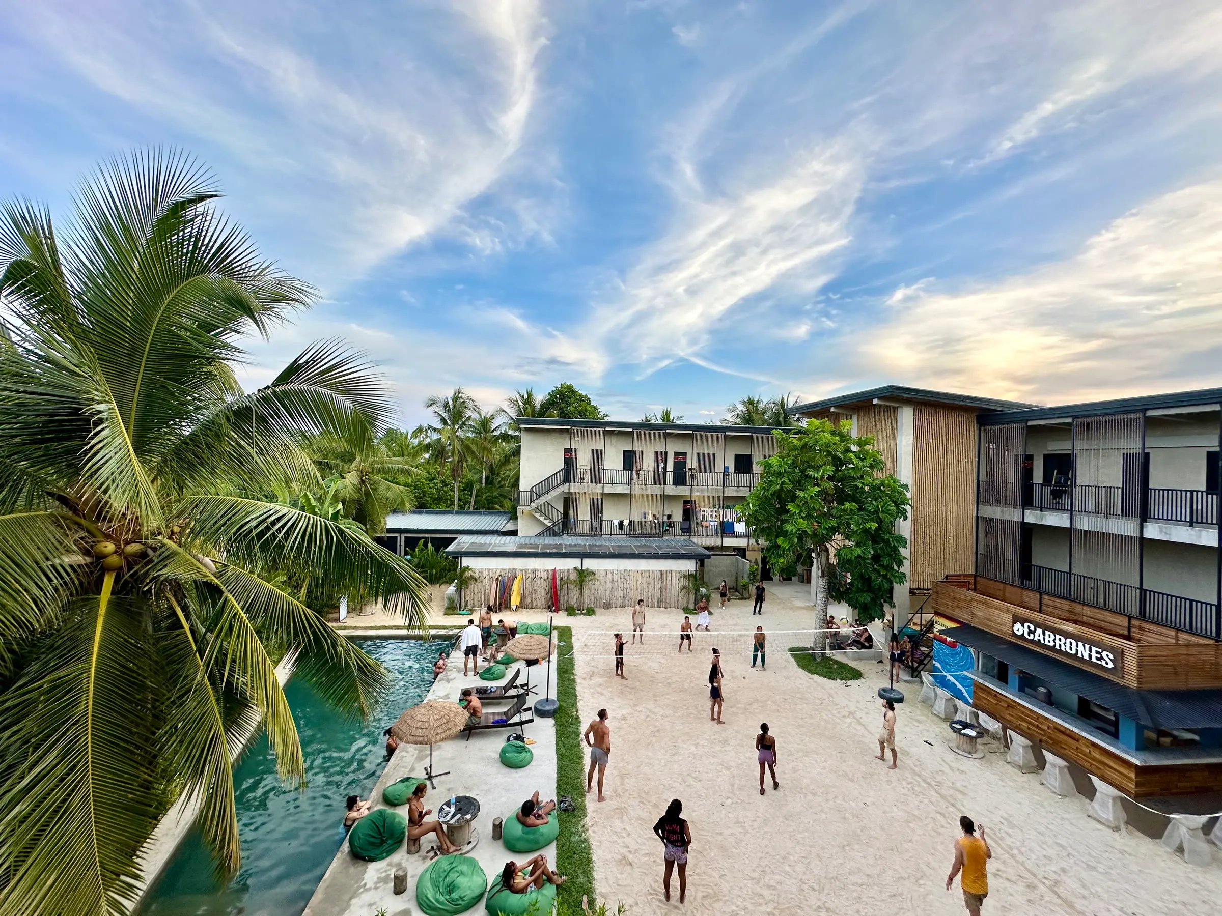 The courtyard of Rucksack Inn Siargao from above: the pool, beanbags and a sandy volleyball court between the two-storey wings, with palms and a blue sky.