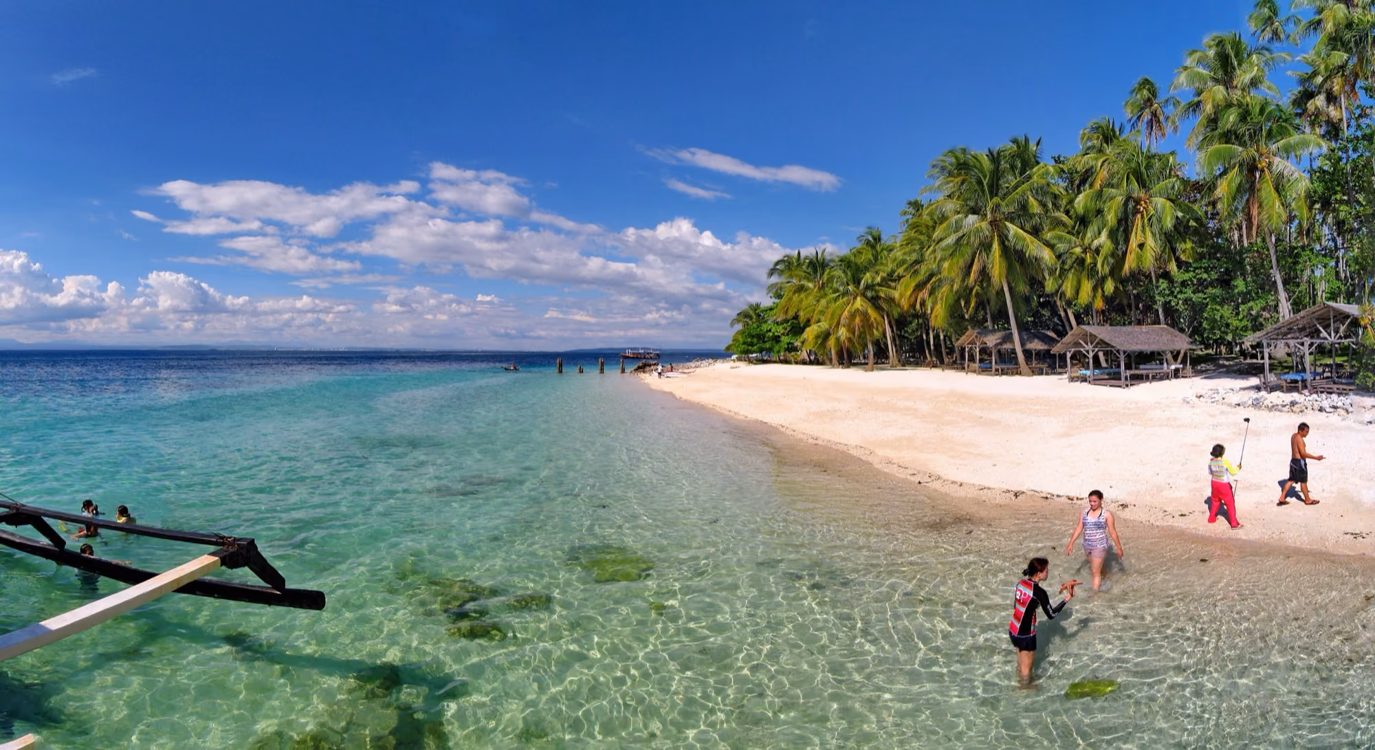 A white sand beach on Samal Island with palm trees, turquoise shallow water and an outrigger in the foreground.