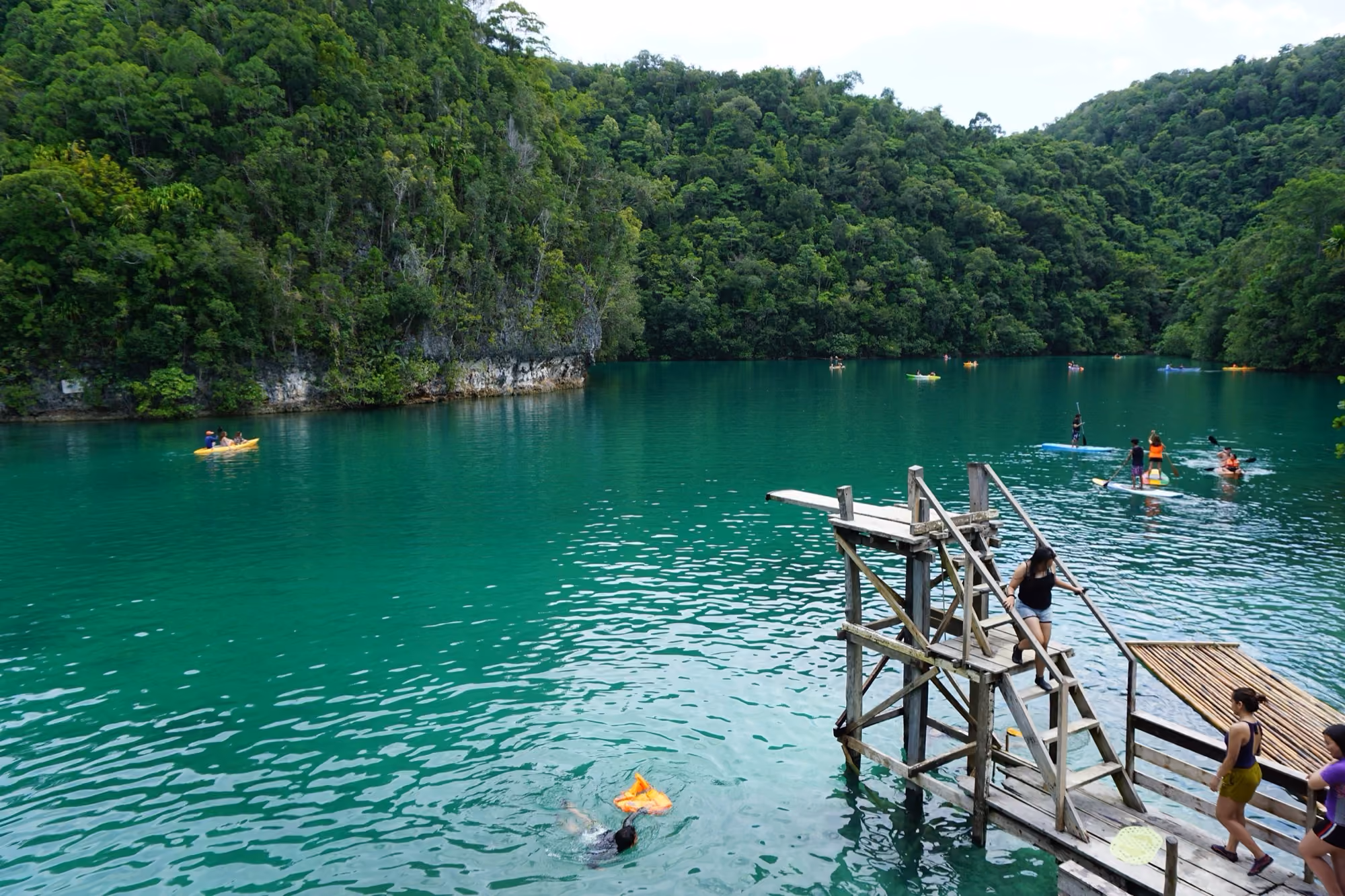 Sugba Lagoon near Siargao: green water ringed by limestone cliffs and jungle, with a wooden jump platform, kayaks and paddleboarders out on the water.