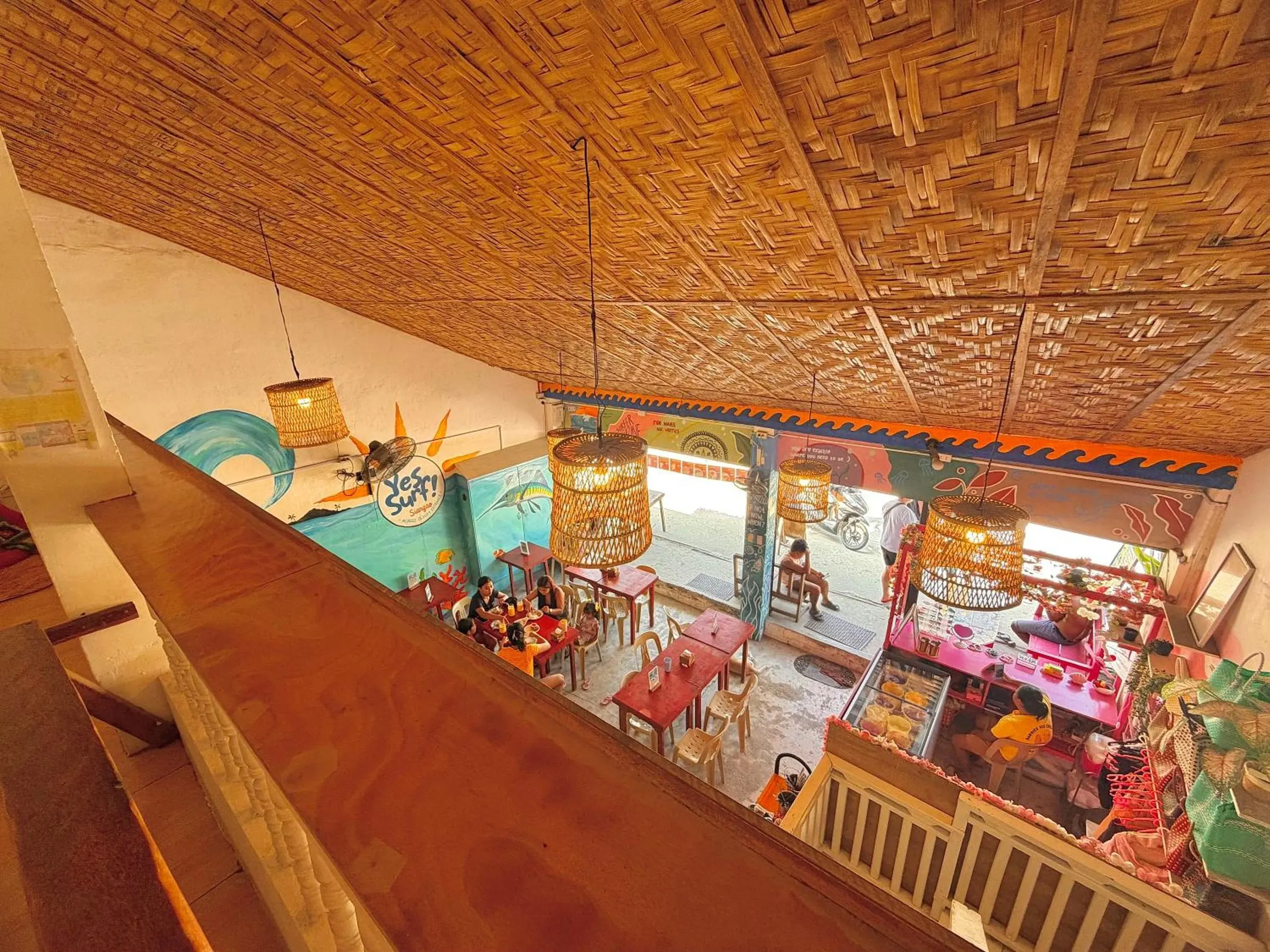 The colourful ground floor at Yes Surf Siargao seen from the upstairs gallery, under a woven bamboo ceiling with rattan lamps.